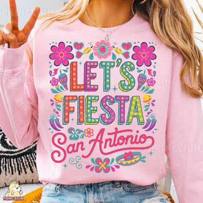 a woman wearing a pink sweatshirt that says let's fiesta san antonio